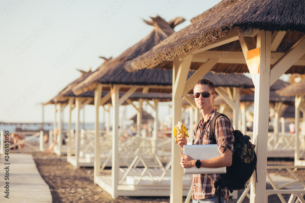 Portrait of a young guy eating a banana with a laptop in his hands standing on a summer canopy of straw. The concept of combining leisure and work in the summer