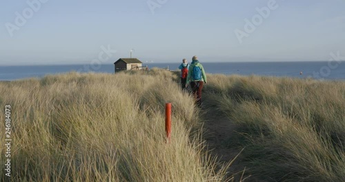 People at the seaside. Tall Grass at the British beach. Tourists walking on the sand dunes towards bird hide. Walney Island, UK. 4K photo