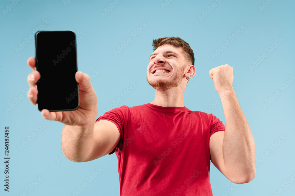 Young handsome man showing smartphone screen over blue background with a surprise face. Human emotions, facial expression concept. Trendy colors Young handsome man showing smartphone screen over blue background with a surprise face. Human emotions, facial expression concept. Trendy colors