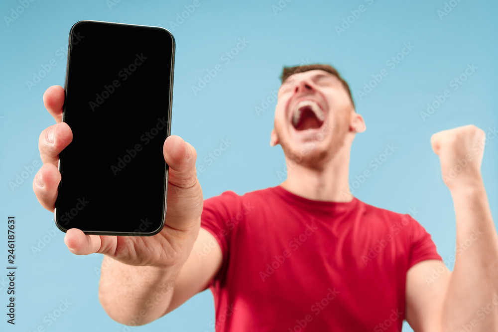 Young handsome man showing smartphone screen over blue background with a surprise face. Human emotions, facial expression concept. Trendy colors - obrazy, fototapety, plakaty 