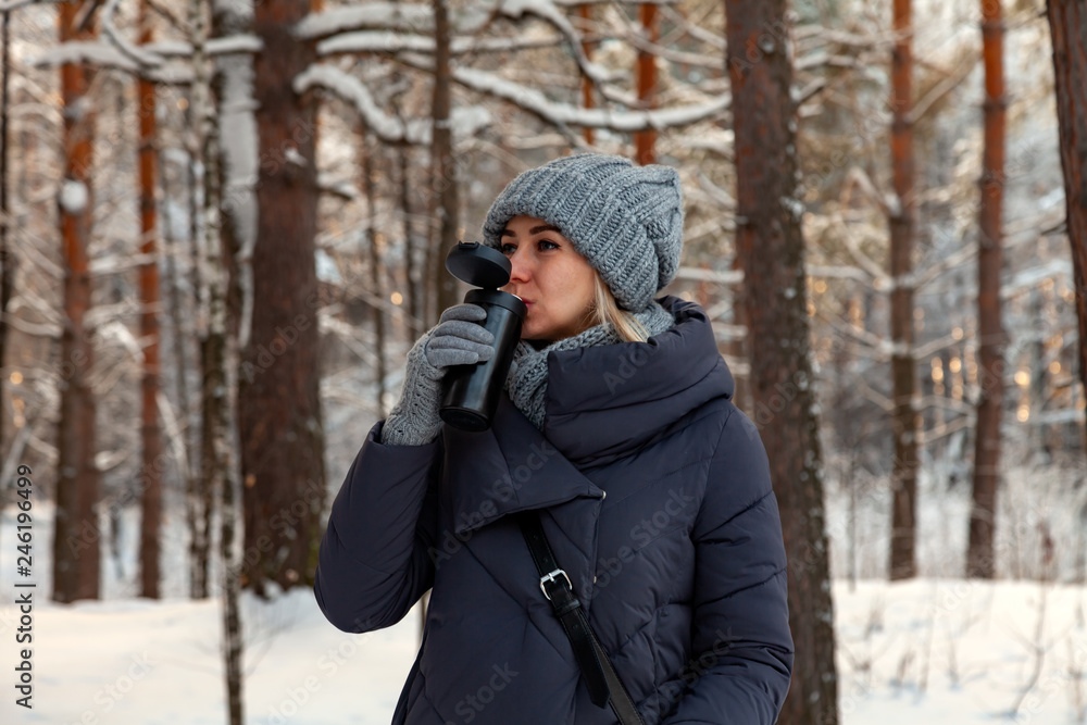 Young beautiful blonde in a knitted hat, scarf and gloves drinks coffee or tea in the winter snow-covered forest at sunset of the day against the backdrop of a lot of trees and sun