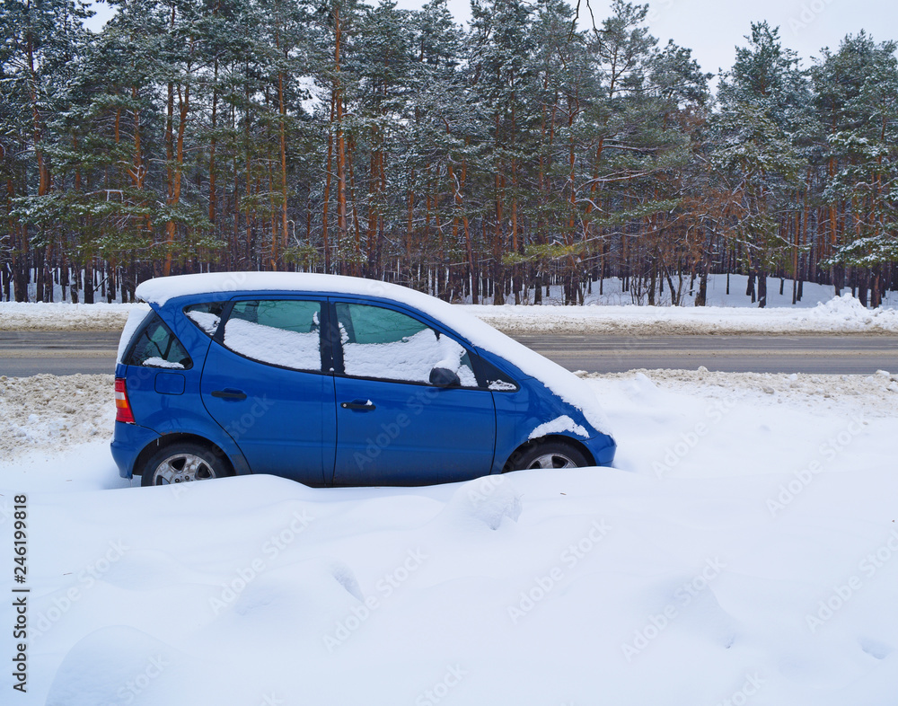 Blue small car in the snow near the road and forest in winter.