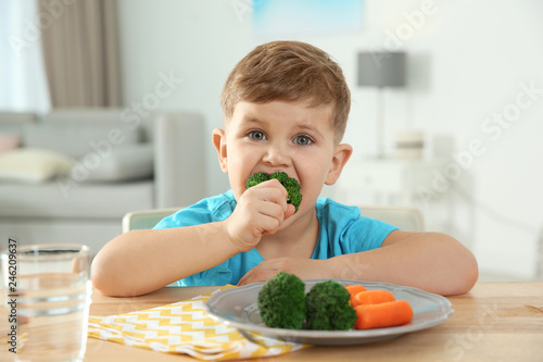 Adorable little boy eating vegetables at table in room