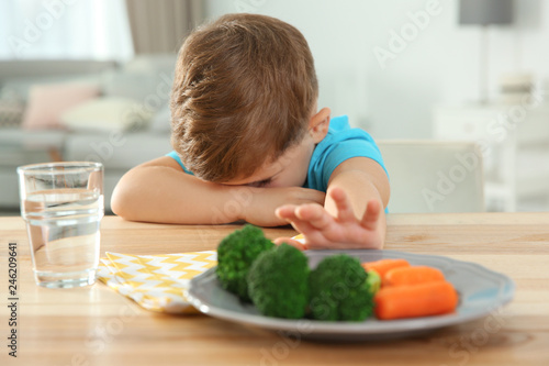 Unhappy little boy refusing to eat vegetables at table in room