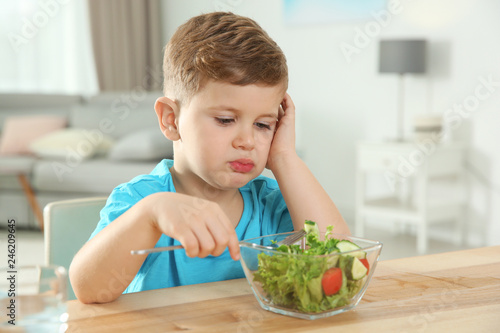 Sad little boy eating vegetable salad at table in room