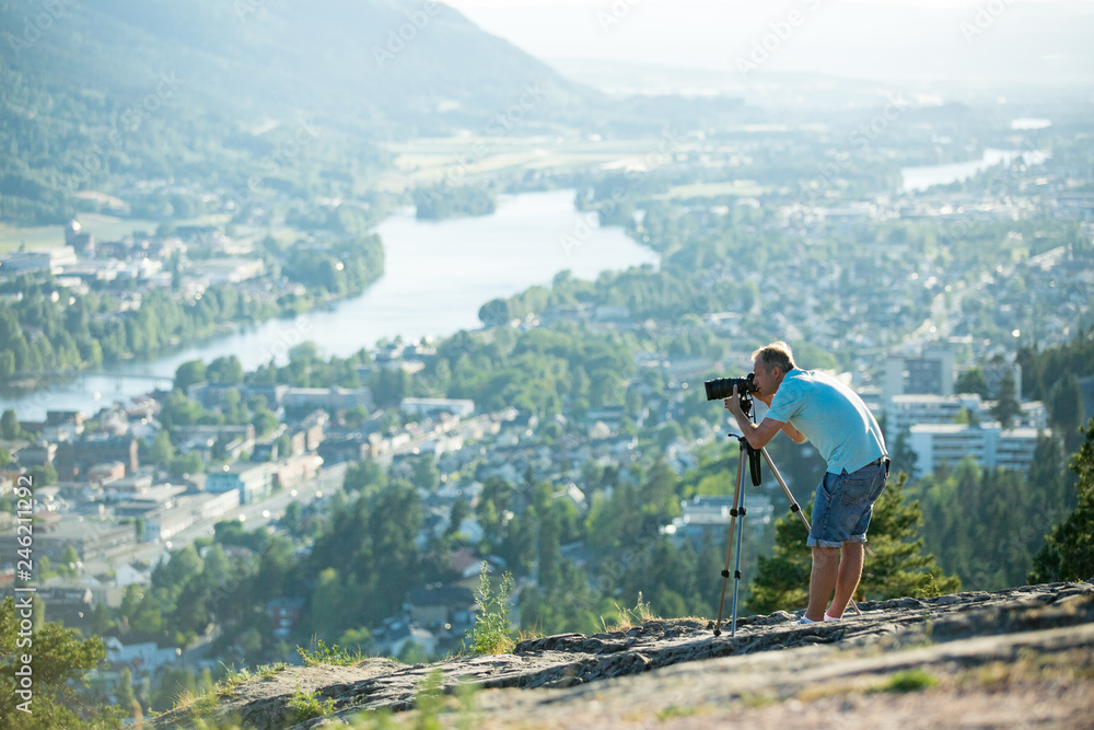Man filming on camera with tripod on the top of mountain. Tourist hiking and taking pictures. Panoramic top view of Drammen city, mountains in fog and valley with river, sunny weather. Norway