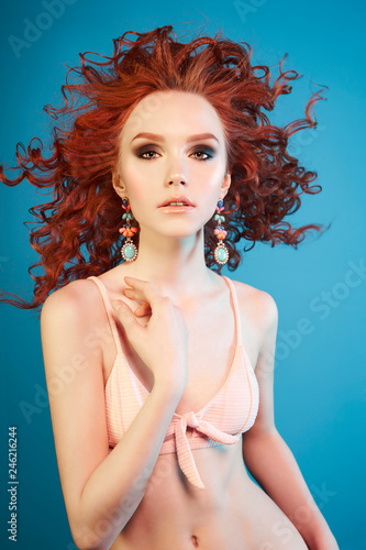 Beautiful sexy woman with red hair and elegant jewelry. Young lady in stylish swimsuit. Modern fashionable makeup with smoky eyes. summer style fashion. Portrait of pretty beauty on blue background.