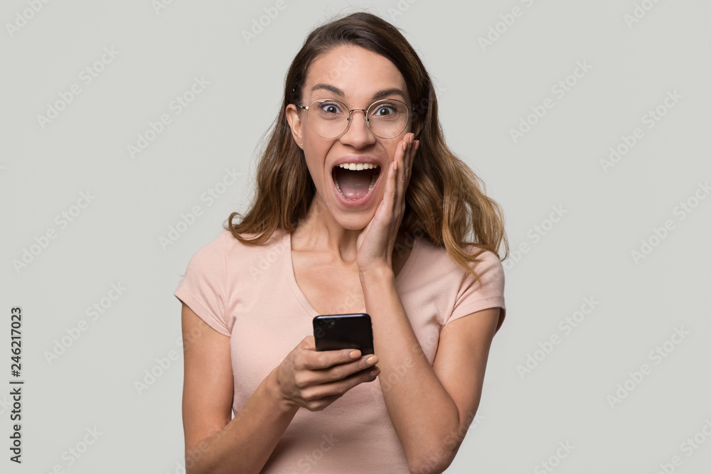 Excited surprised young woman winner amazed by good news holding phone looking at camera, euphoric emotional girl scream with joy happy about mobile win isolated on white grey blank studio background Excited surprised young woman winner amazed by good news holding phone looking at camera, euphoric emotional girl scream with joy happy about mobile win isolated on white grey blank studio background