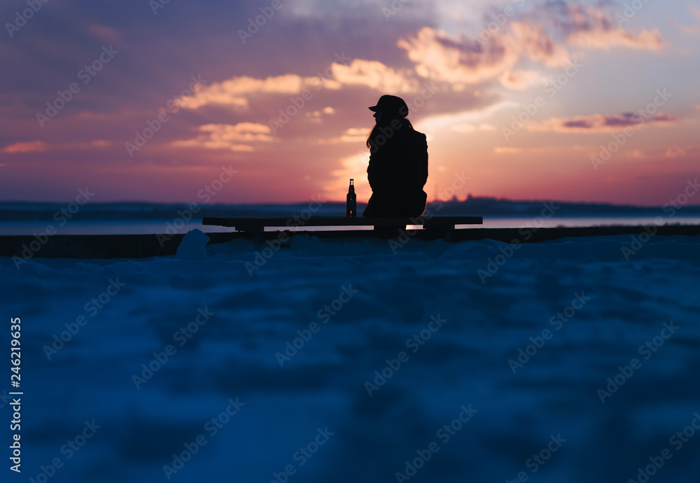 A young girl sits on a bench and drinks alcohol alone. Female alcoholism. Silhouette of a girl with a bottle. Depression.