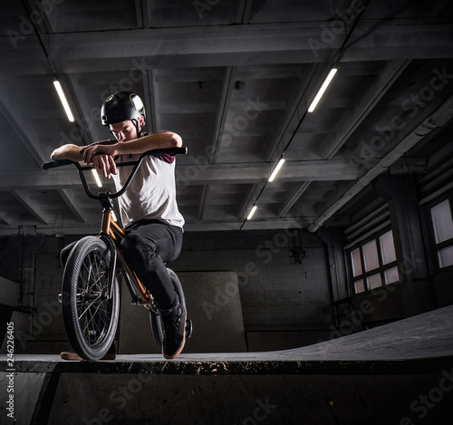 Tired BMX rider in protective helmet sitting on his bicycle in a skatepark indoors