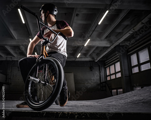 Professional BMX rider in protective helmet sitting on his bicycle in a skatepark indoors
