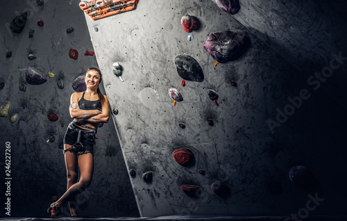 Full body portrait of a cheerful female climber leaning on a bouldering wall at the gym.