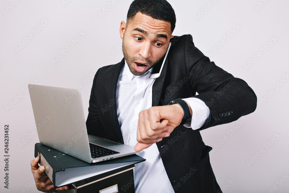 Busy handsome guy in white shirt and black jacket talking on phone, holding folders, laptop, looks astonished at watch on white background. Being late, meeting, office worker, career Busy handsome guy in white shirt and black jacket talking on phone, holding folders, laptop, looks astonished at watch on white background. Being late, meeting, office worker, career