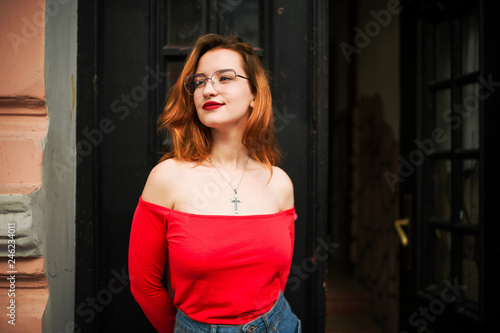 Attractive redhaired woman in eyeglasses, wear on red blouse and jeans skirt posing at street against old wooden door.