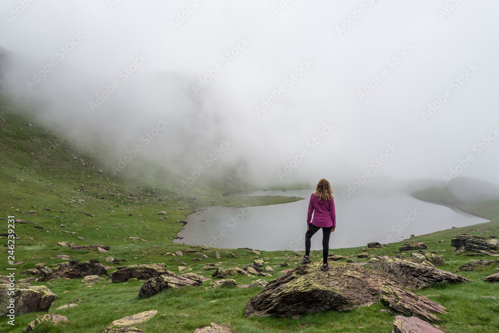 A young woman observes a high mountain lake in the Pyrenees, in Spain, on a foggy day.