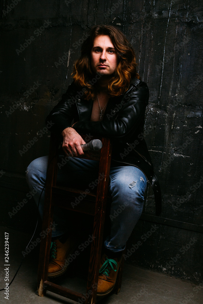 a portrait of a brutal, handsome man, a singer who sits on a chair on a dark background, in a black leather kurt on his naked body and holds a microphone in his hands. presenter, singer at the concert a portrait of a brutal, handsome man, a singer who sits on a chair on a dark background, in a black leather kurt on his naked body and holds a microphone in his hands. presenter, singer at the concert