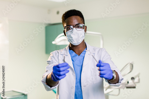 Portrait of attractive handsome black dentist in protective face mask, white lab coat, blue tie, holding equipments in his arms