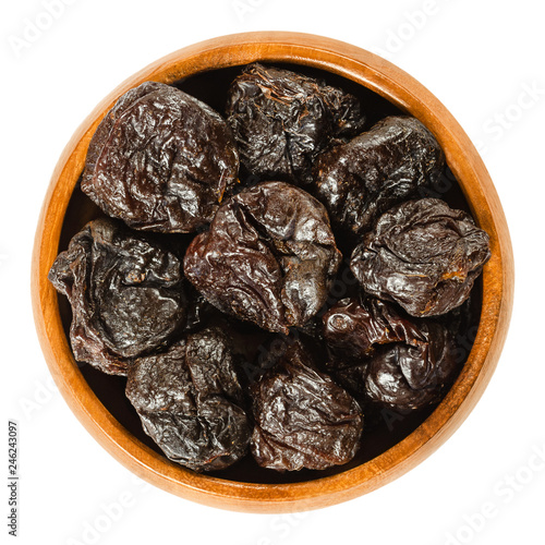 Prunes, dried plums in wooden bowl. Uncooked, dehydrated, pitted fruits of Prunus domestica with black color, used as snack. Isolated macro food photo, closeup, from above, on white background.