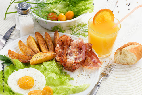 Homemade breakfast with potatoes with crispy bacon, tender egg and a glass of fresh juice on a white table background.