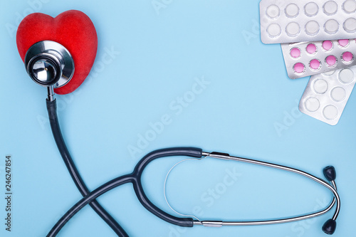 Blue background lies a statoscope, a red saddle and pills. The concept of treatment of cardiovascular diseases. Closeup of painted heart, lot of drugs and stethoscope on blue background.