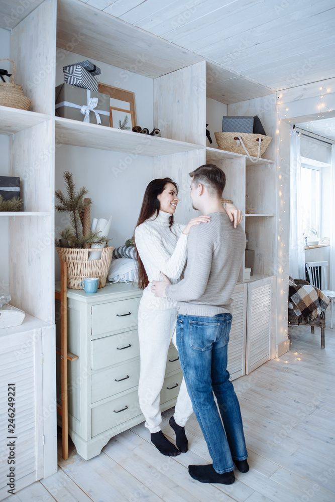 Young caucasian couple sitting at home in the living room in Scandinavian style, hugging and smiling. Young lovers having good time. A man and a woman dressed in the bright winter sweater Young caucasian couple sitting at home in the living room in Scandinavian style, hugging and smiling. Young lovers having good time. A man and a woman dressed in the bright winter sweater