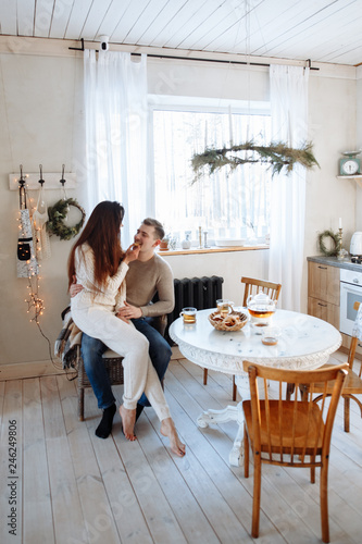 Young caucasian couple sitting at home kitchen, hugging, smiling, drinking tea. Young lovers having good time. Man and woman communicate sitting at a table in the dining room.Winter decor.