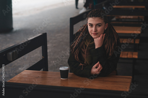 stylish portrait of an attractive woman with long pigtails, in an eco-coat, scarf, sitting at a table outdoors holding coffee cup of hot coffee