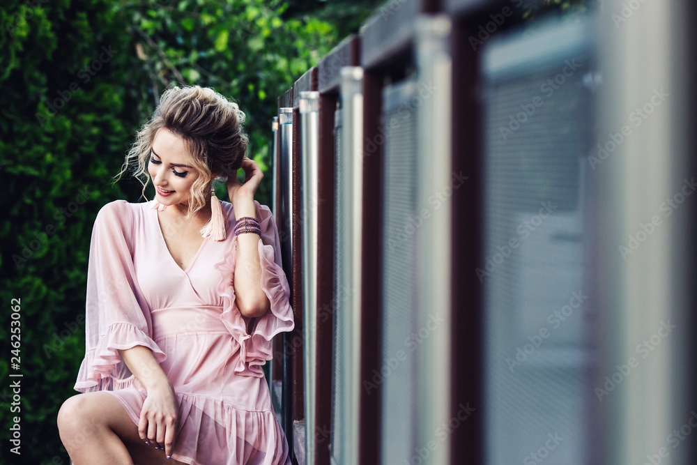 attractive young girl in a short pink dress posing near a steel fence on the background of green foliage. attractive young girl in a short pink dress posing near a steel fence on the background of green foliage.