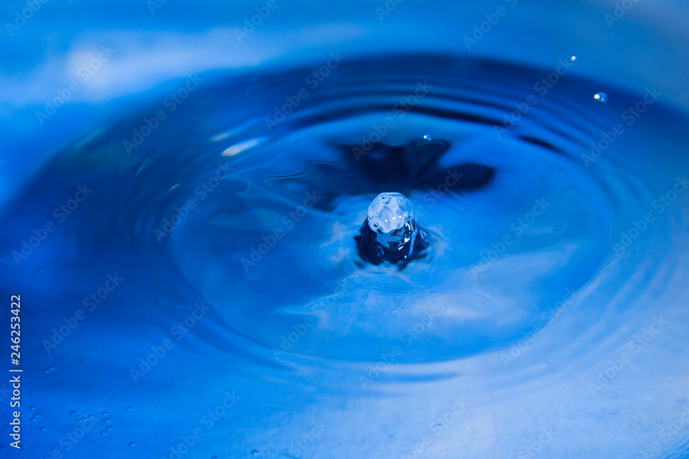 Water droplet falls into the blue water and creates small column close up.