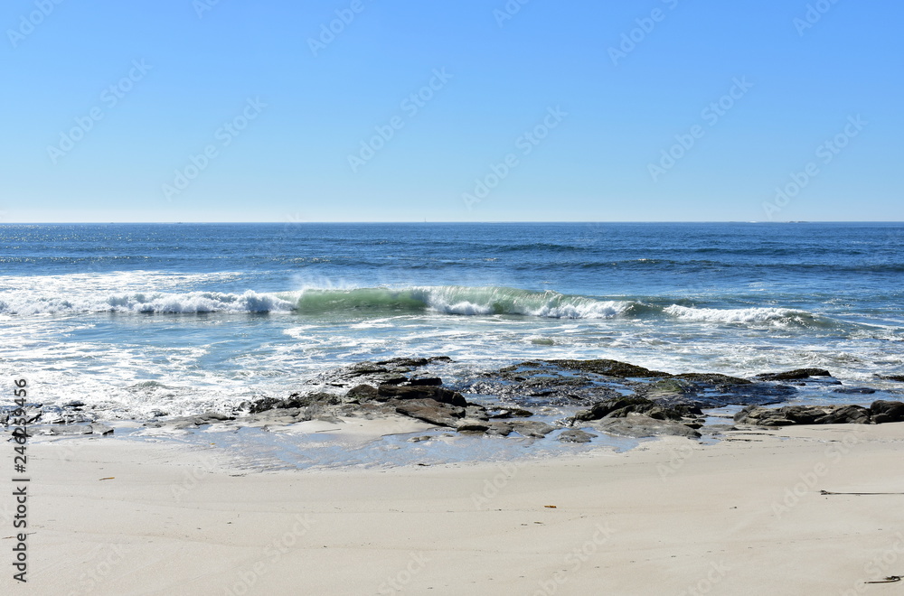Beach with rocks, bright sand and small waves breaking. Blue sea with foam, sunny day. Galicia, Spain.