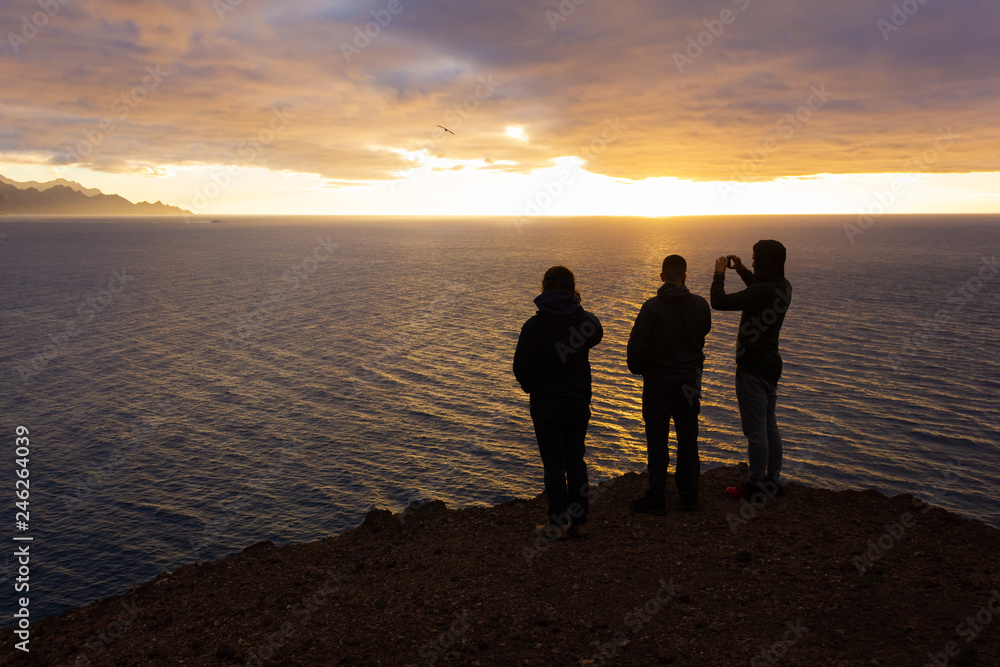Silhouettes of three men standing on cliff edge enjoying sunset by the sea in Gran Canaria, Spain. Group of friends taking photos of splendid twilight with cellphone in Canary Islands. Travel concept