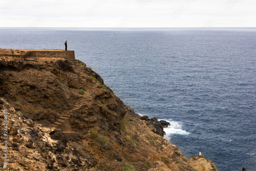 Man standing on cliff edge by the sea. Person alone on top of mountain facing the ocean on cloudy day. Loneliness, solitude, depression concepts
