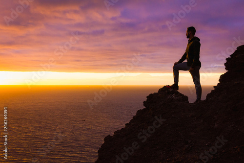 Visionary man standing on top of cliff edge staring at colorful sunset by the sea in Gran Canaria. Silhouette of person witnessing unique twilight from mountain top. Successful  entrepreneur concepts