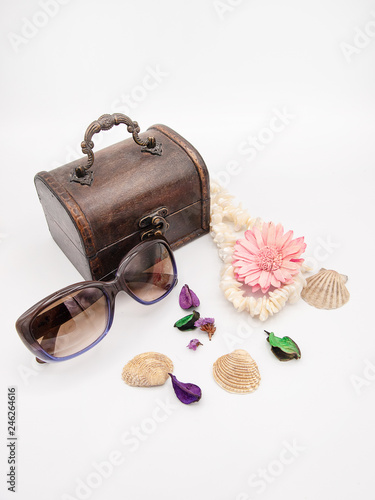 Travel or vacation concept with a wooden treasury chest jewelry box, stylish sunglasses, sea pearls necklace, sea shells, rocks, flower petals and a decorative chrysanthemum flower isolated on white