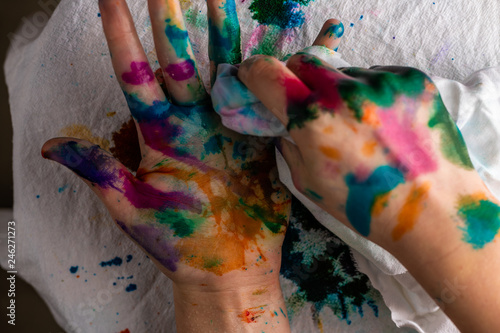 A child cleans up hands that are covered in red, pink, yellow, orange, red, blue, green, and purple ink. Concepts: art, education, play, watercolor, finger painting, mess, creativity, fun, enjoyment,