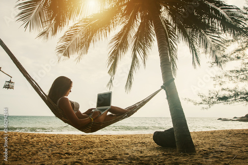 Young woman working on laptop seating in hammock under palm trees on tropical beach