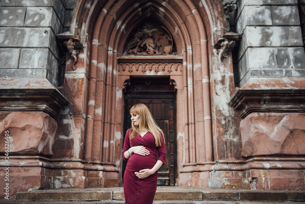 Pregnant girl blonde in burgundy dress. In the big city. Long hair. 9 months waiting. Happiness to be a mother. Against the backdrop of a beautiful old building.