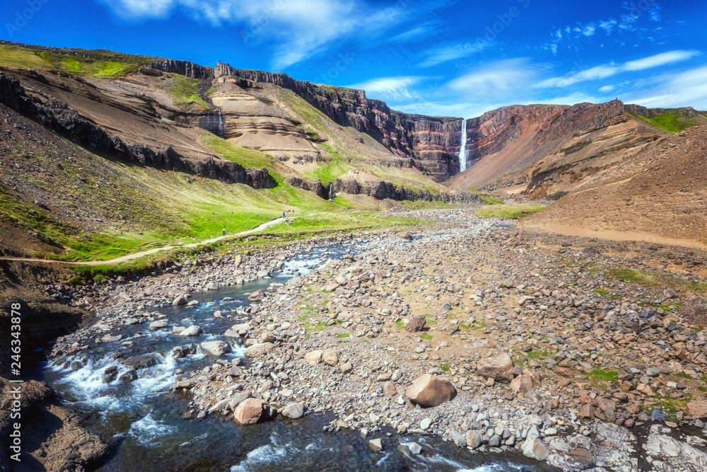 Amazing nature, scenic view of Hengifoss waterfall in Iceland falling into a magnificent gorge, colorful rock and rugged river. Icelandic summer landscape with blue sky, outdoor travel background
