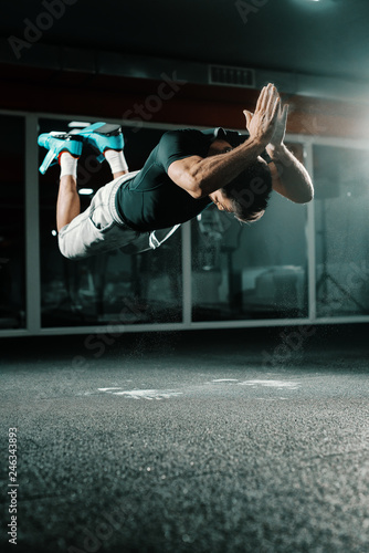 Muscular Caucasian bodybuilder doing jumping push ups in dark. Dust in the air, gym interior, mirror in background.