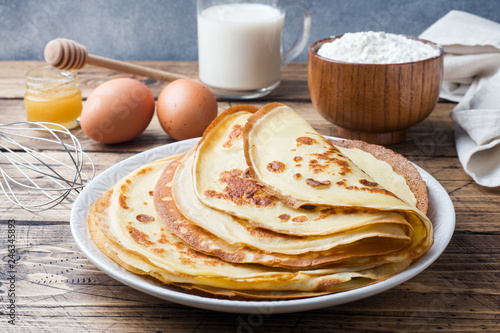 Thin pancakes on a plate. Wooden background. Ingredients to cooking eggs, milk, flour.