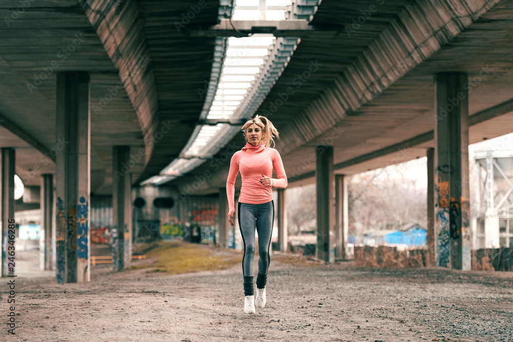 Gorgeous blonde woman wearing sportswear and with ponytail running under the bridge.
