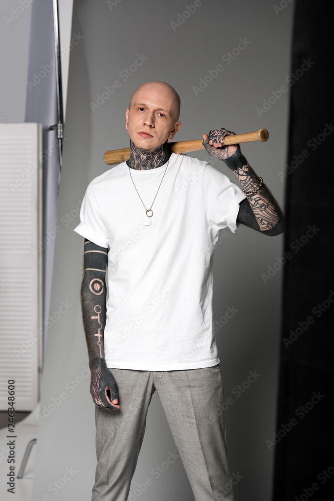 bald tattooed man in white t-shirt standing with baseball bat on shoulder  and looking at camera in studio Stock Photo | Adobe Stock