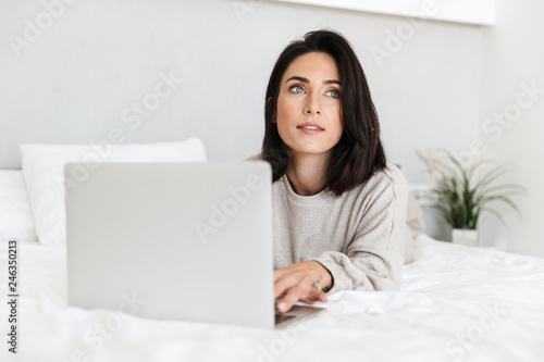 Photo of satisfied woman 30s using laptop, while lying in bed with white linen in bright room