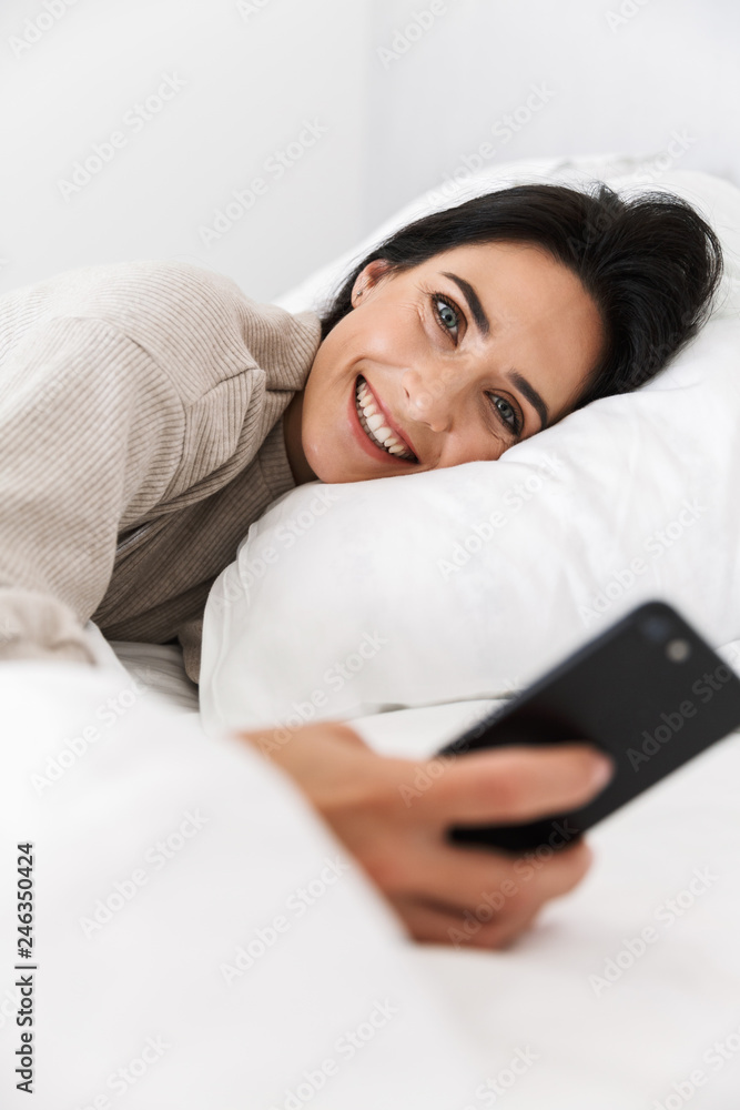 Photo of joyous woman 30s using smartphone, while lying in bed with white linen at home Photo of joyous woman 30s using smartphone, while lying in bed with white linen at home