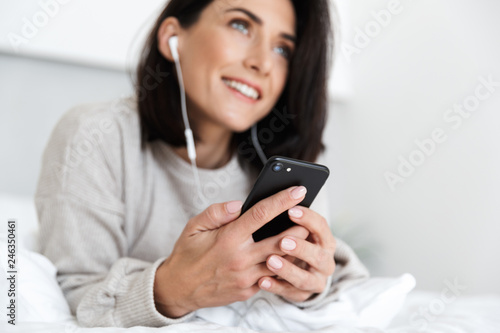 Image of caucasian woman 30s wearing earphones holding cell phone, while lying in bed in bright room