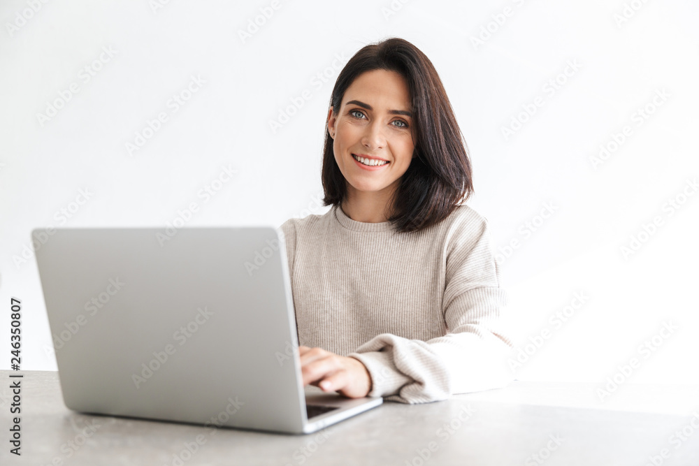 Image of adorable woman 30s working on laptop, while sitting over white wall in bright room Image of adorable woman 30s working on laptop, while sitting over white wall in bright room