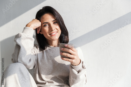 Image of happy woman 30s holding cup with tea, while sitting over white wall indoor photo