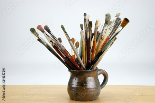 A collection of well used art paint brushes in an old jug in front of a white background