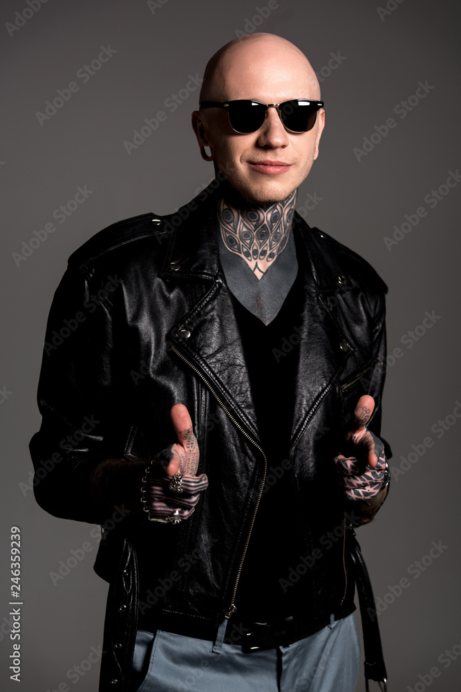 Obraz premium smiling tattooed man in leather jacket and sunglasses pointing at camera isolated on grey
