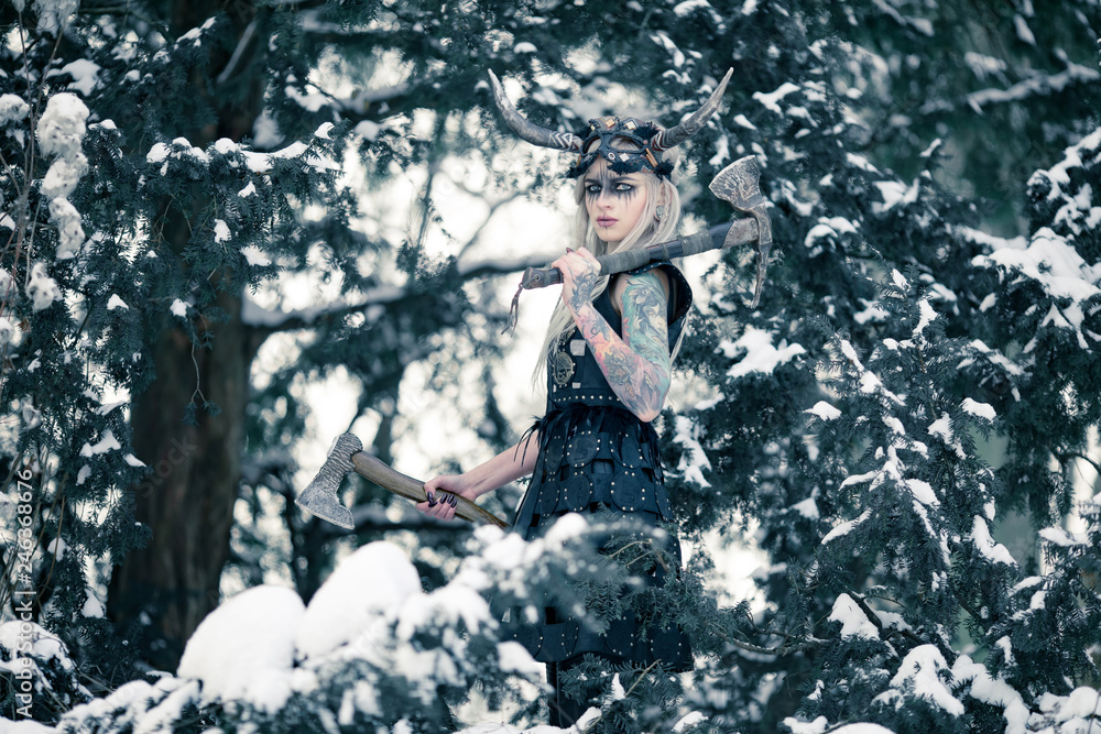 Beautiful warrior woman in image of viking with horned helmet and two axes in winter snowy forest. Beautiful warrior woman in image of viking with horned helmet and two axes in winter snowy forest.